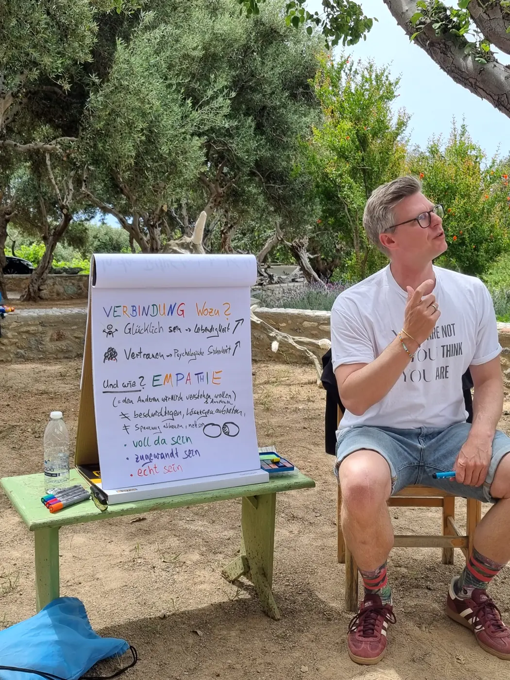 Ein Trainer sitzt neben einem Flipchart zum Thema Verbindung und Empathie, unter Olivenbäumen