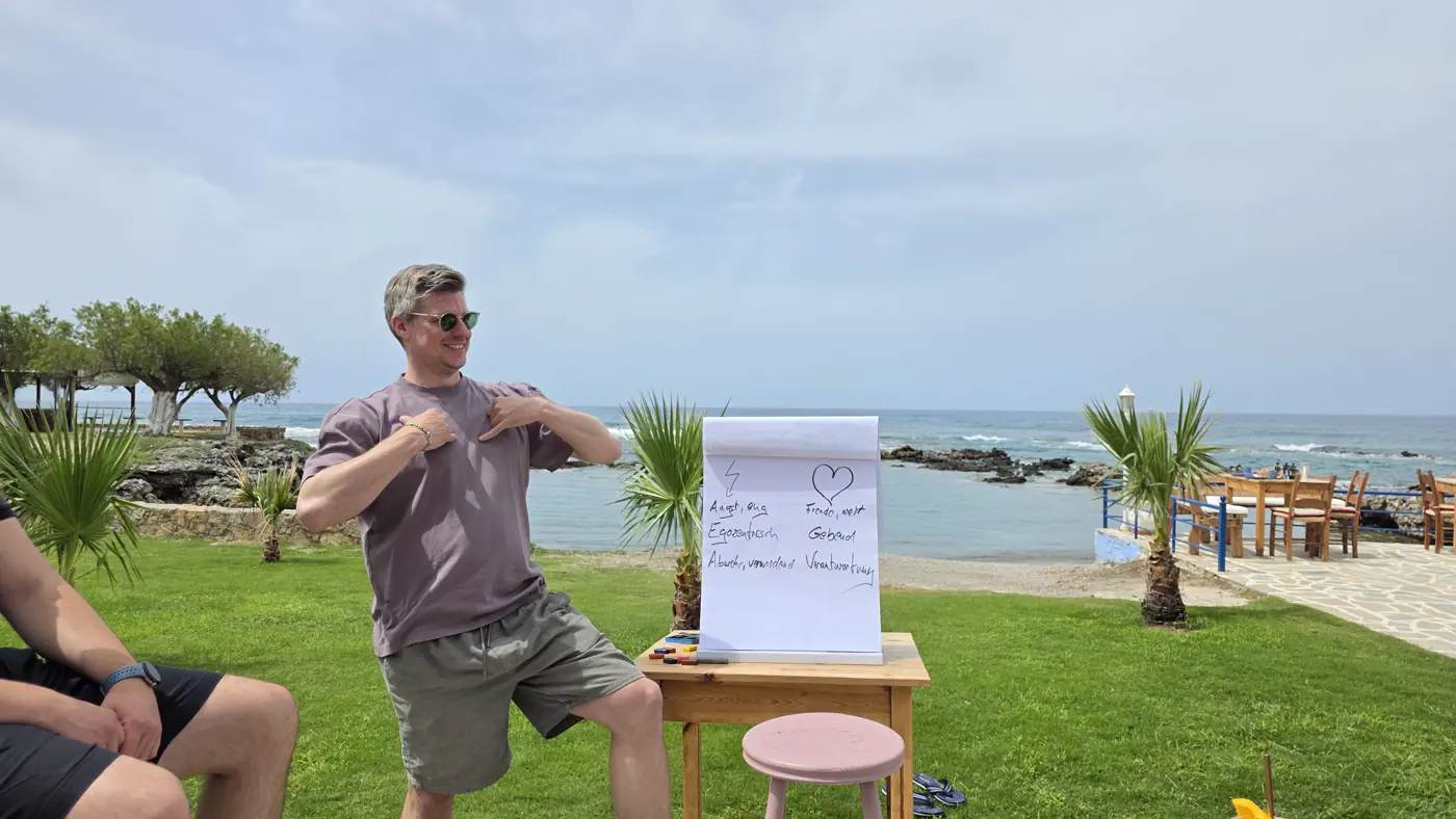 Ein Teilnehmer steht lachend neben einem Flipchart auf einer Wiese am Meer und zeigt auf sich selbst