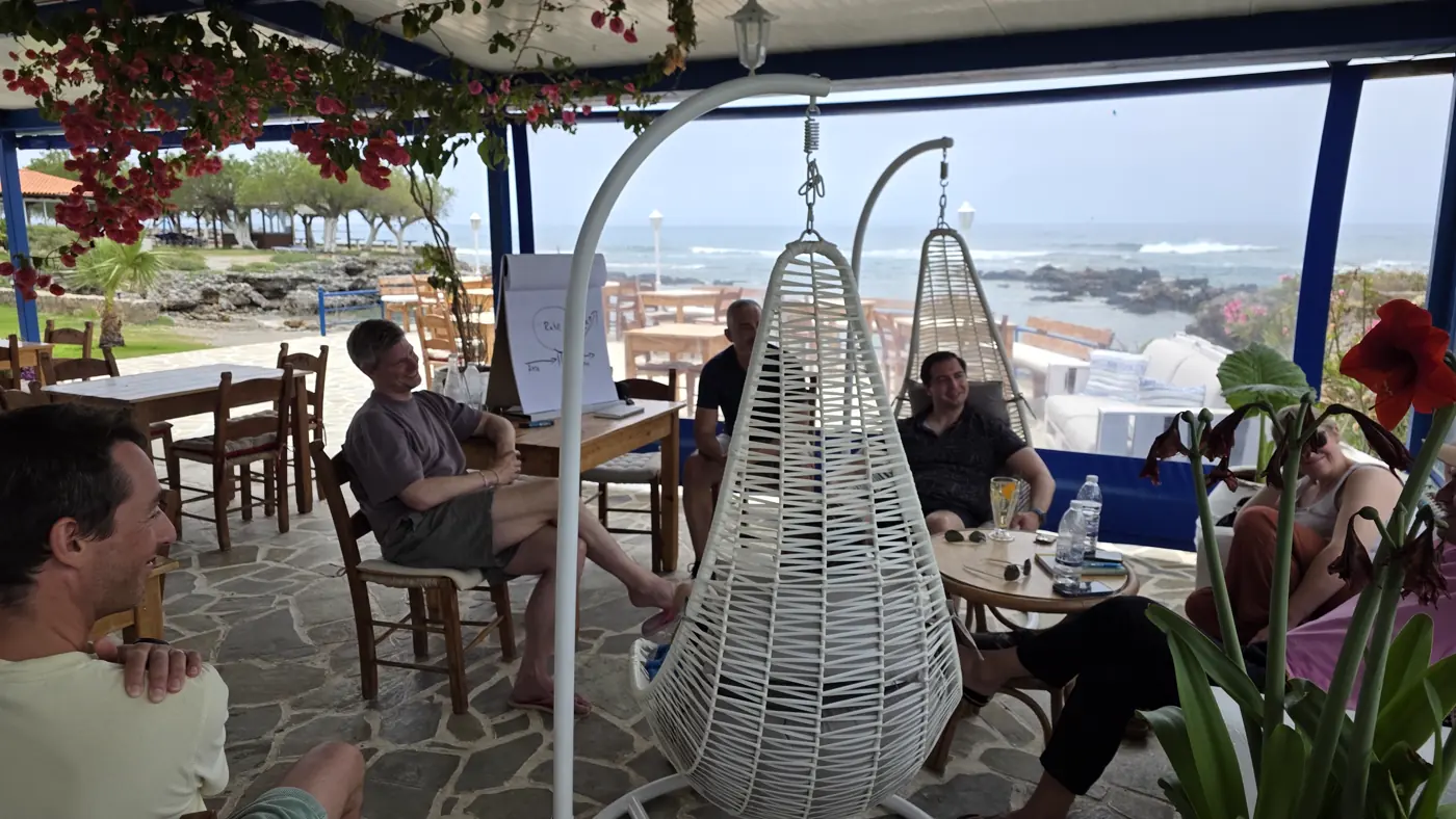 Die Gruppe sitzt unter einem überdachten Pavillon mit Hängesesseln und Flipchart, dahinter das Meer