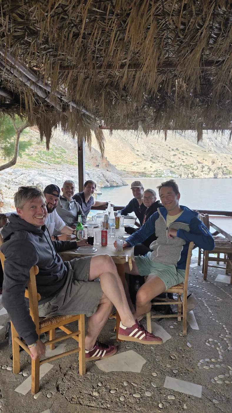 Die Gruppe am Tisch einer Taverne unter einem Schilfdach, dahinter eine Bucht und steile Felsen