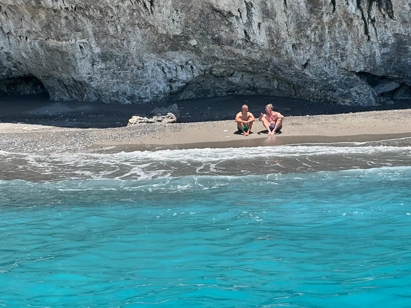 Zwei Teilnehmer sitzen im Gespräch an einem kleinen Strand unter hellen Felsen, vorne türkisblaues Wasser