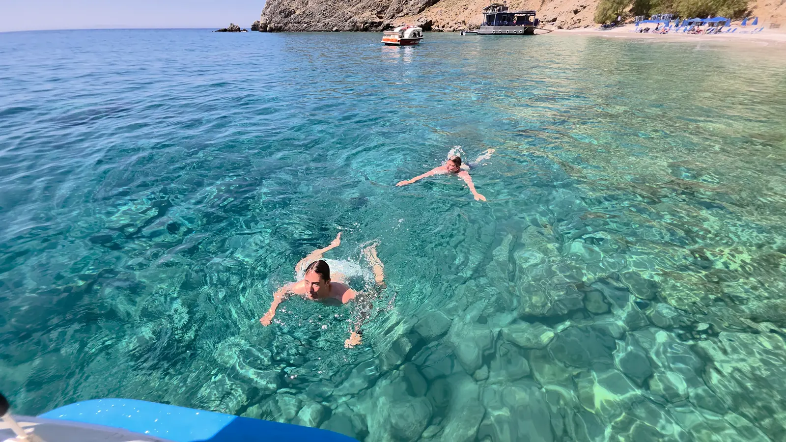 Zwei Teilnehmer schwimmen im glasklaren, türkisblauen Wasser einer Bucht, dahinter ein Boot und ein Strand